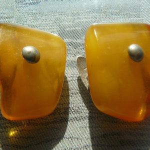 May include: A pair of amber clip-on earrings with silver accents. The earrings are shaped like a rectangle with rounded edges.