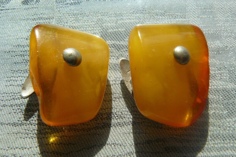 May include: A pair of amber clip-on earrings with silver accents. The earrings are shaped like a rectangle with rounded edges.