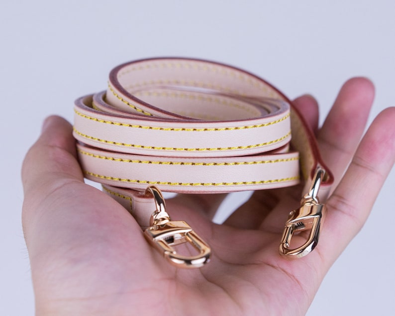 May include: A beige leather strap with gold-tone hardware. The strap is folded in half and has a double row of stitching.