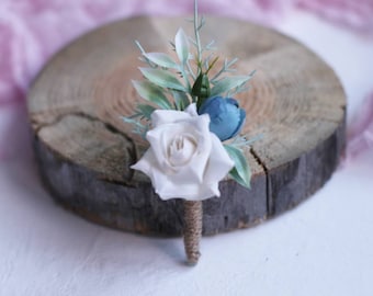 Puede incluir: Una rosa blanca con flores azules y hojas verdes, arreglada en un boutonniere.