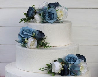 May include: A three-tiered white wedding cake decorated with blue and white artificial flowers. The cake is on a white surface.