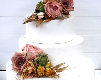 May include: A three-tiered white wedding cake decorated with blush pink, yellow, and brown floral arrangements. The cake is adorned with gold accents.