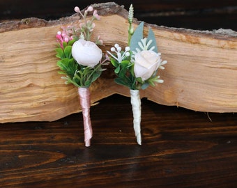 Puede incluir: Dos pequeños boutonnières florales con flores blancas y vegetación. Un boutonnière tiene acentos rosados ​​y una cinta rosa, el otro tiene una cinta blanca.