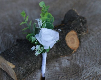 Puede incluir: Un boutonniere de rosa blanca con vegetación y una cinta blanca.