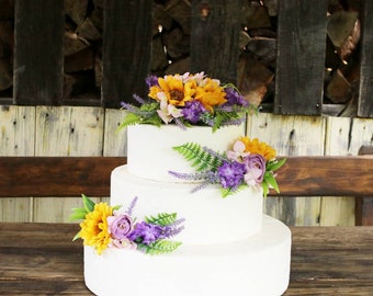 May include: A three-tiered white wedding cake decorated with yellow sunflowers, purple lavender, and pink flowers. The cake is on a wooden table.