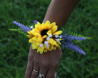 May include: A yellow sunflower with white accents and purple lavender flowers are arranged in a wrist corsage.