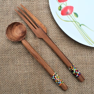 Puede incluir: Juego de cuchara y tenedor de madera para servir ensalada con mangos con cuentas. La cuchara tiene un cuenco redondo y el tenedor tiene tres púas. Los mangos están adornados con cuentas de colores. Un plato blanco con arte floral rojo está en el fondo.