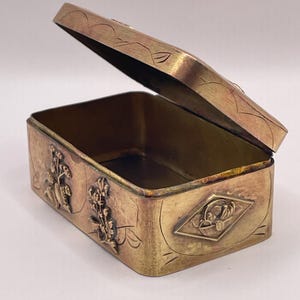 Puede incluir: Caja rectangular de latón antiguo con tapa abisagrada ligeramente abierta. La caja presenta diseños florales y geométricos en relieve en los laterales y la tapa. El interior es de latón más oscuro.
