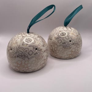 Op de afbeelding: Twee bolvormige, crèmekleurige keramische zout- en peperstrooiers met een gouden bloemenpatroon. Elke strooier heeft een teal lintlus om op te hangen. De strooiers hebben twee gaatjes om te doseren.