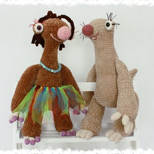 May include: Two crocheted stuffed animals, one brown with a colorful tutu and the other beige, sitting on a white bench.