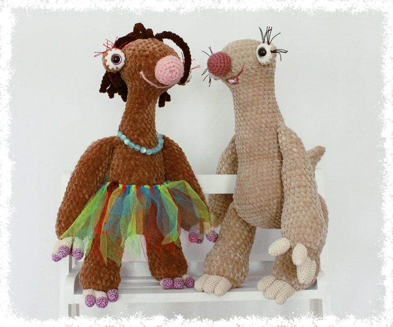 May include: Two crocheted stuffed animals, one brown with a colorful tutu and the other beige, sitting on a white bench.