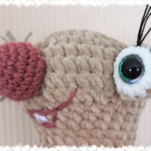 May include: Close-up of a crocheted plush toy with a brown body, a pink nose, and a green glitter eye. The toy has black yarn whiskers.
