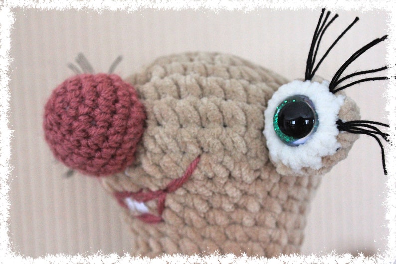 May include: Close-up of a crocheted plush toy with a brown body, a pink nose, and a green glitter eye. The toy has black yarn whiskers.
