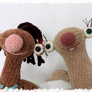 May include: Two crocheted plush toys with large eyes and pink noses. The brown toy has brown hair and is wearing a turquoise beaded necklace. The beige toy has black eyelashes.
