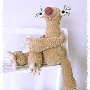May include: A crocheted sloth plush toy with a brown and beige fur, sitting on a white shelf. The sloth has a pink nose, green eyes, and a big smile.