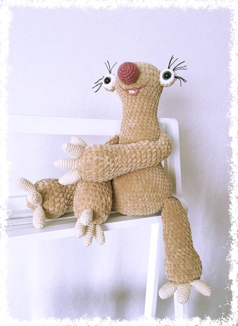 May include: A crocheted sloth plush toy with a brown and beige fur, sitting on a white shelf. The sloth has a pink nose, green eyes, and a big smile.