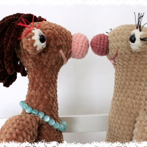 May include: Two crocheted stuffed animals, one brown with a blue beaded necklace and a pink nose, the other tan with a pink nose, facing each other.