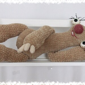 May include: A crocheted plush toy in the shape of a sloth, lying on a white shelf. The sloth has brown fur, a pink nose, and black eyelashes.