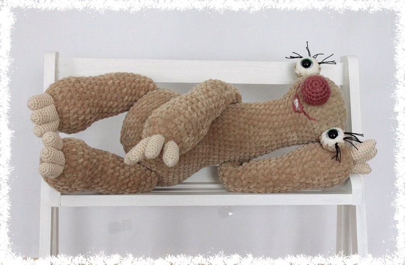 May include: A crocheted plush toy in the shape of a sloth, lying on a white shelf. The sloth has brown fur, a pink nose, and black eyelashes.