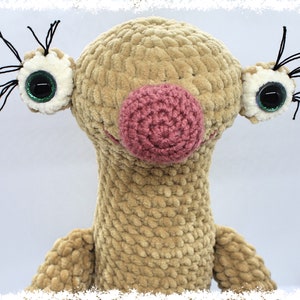 May include: A crocheted plush toy with a brown body, pink nose, and large green eyes. The toy has black yarn for eyelashes.