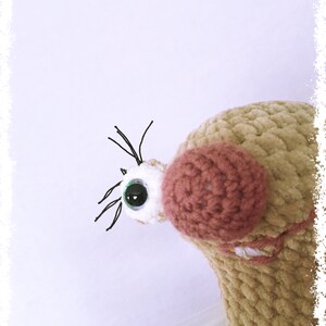 May include: A close-up of a crocheted plush toy with a brown and pink body, a white eye with a green pupil, and black eyelashes. The toy is looking to the right.