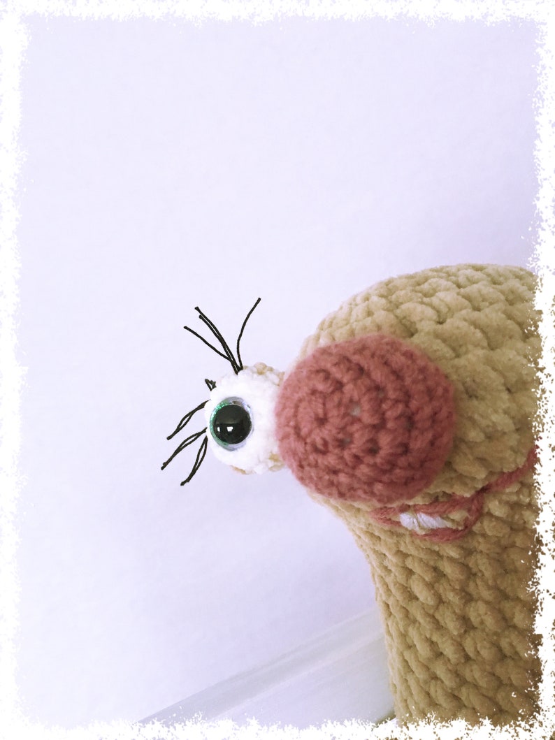 May include: A close-up of a crocheted plush toy with a brown and pink body, a white eye with a green pupil, and black eyelashes. The toy is looking to the right.
