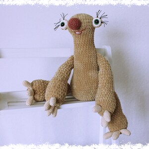May include: A crocheted sloth plush toy with brown fur, large green eyes, a pink nose, and a wide smile. The sloth is sitting on a white shelf.
