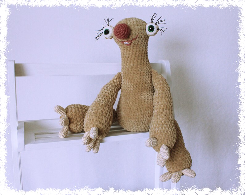May include: A crocheted sloth plush toy with brown fur, large green eyes, a pink nose, and a wide smile. The sloth is sitting on a white shelf.