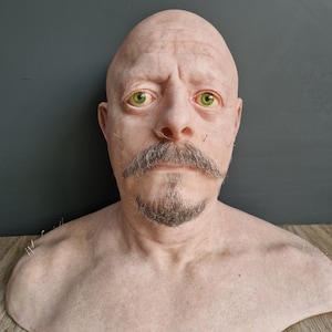 Puede incluir: Un busto realista de un hombre calvo con barba y bigote. El busto está hecho de un material de color carne y tiene detalles realistas, incluyendo arrugas y poros.