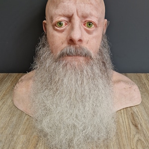 May include: A close-up of a bald man's face with a long, white beard. The man has green eyes and a serious expression. The beard is very detailed and realistic, with individual hairs visible. The man's face is also very detailed, with wrinkles and lines visible. The background is a dark gray.