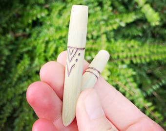 May include: Two light-colored wooden dowels with etched designs are held in a hand. One dowel has a chevron pattern, and the other has two horizontal lines. The background is a blurred green foliage.