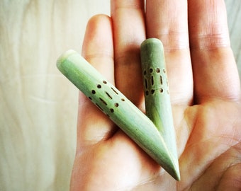 May include: Two light green wooden dowels with decorative cut-out patterns. The dowels are held in a hand, and have a cylindrical shape with a pointed end. The dowels are approximately 5 inches long.