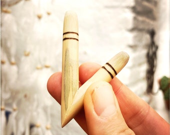 May include: Two light-colored wooden objects with a pointed end and a rounded top, each featuring two dark brown bands. The objects are held in a hand against a blurred, white background, possibly a dreamcatcher.