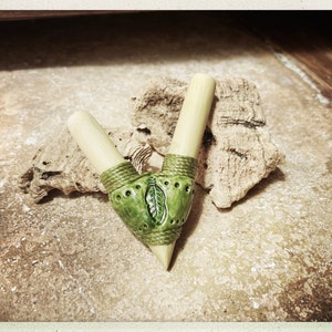 Peut inclure: Une pipe à fumer artisanale en forme de V. Elle se compose de deux tiges en bambou clair reliées à un foyer en céramique verte. Le foyer est orné d'un motif de feuille et entouré de ficelle verte. La pipe repose sur une surface texturée brun clair.