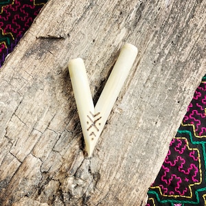 Può includere: Un oggetto di legno a forma di V, di colore chiaro, con un disegno decorativo inciso. L'oggetto è posto su una superficie di legno stagionato, con un tessuto fantasia colorato sullo sfondo. Il disegno è un simbolo.