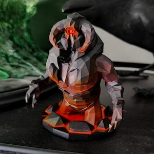 Puede incluir: Escultura geométrica detallada de un demonio con cuernos y ojos rojos brillantes y acentos de fuego. La figura es principalmente gris oscuro y marrón, con una base de naranja y rojo vibrantes. Una estatua de cuervo negro está en el fondo.