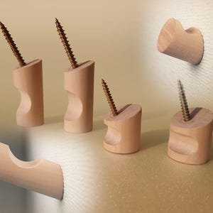 Puede incluir: Una colección de ganchos de pared de madera de color claro, cada uno con una forma cilíndrica y un recorte cóncavo. Algunos ganchos se muestran con un tornillo de metal que sobresale de la parte superior, mientras que otros están montados sobre una superficie texturizada.