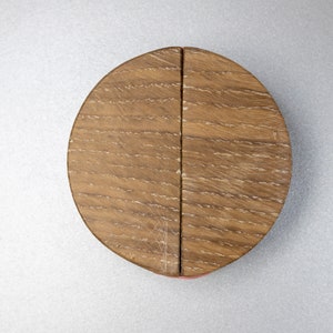 May include: A circular wooden lid with a visible grain pattern, divided into two equal halves by a straight vertical seam. The wood features a medium brown tone with lighter highlights along the textured surface of the circular object.