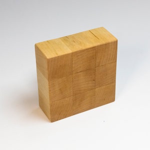 May include: A rectangular block made of light-colored natural wood, featuring a grid pattern of nine square sections on its front face. The object has a smooth, unfinished surface with visible wood grain patterns and straight, clean edges.