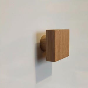 Op de afbeelding: Een houten wandhaak met een vierkant ontwerp. De haak is gemaakt van lichtbruin hout en heeft een cilindrische basis. Het vierkante deel van de haak is met de cilindrische basis aan de muur bevestigd.