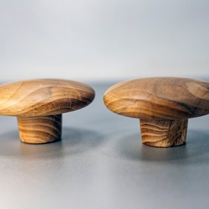 Puede incluir: Dos tiradores de armario de madera con forma de hongo. Los tiradores son de madera de color marrón claro con vetas visibles. Cada tirador tiene una parte superior redondeada y una base cilíndrica, ideal para herrajes de muebles.