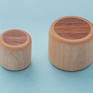 Puede incluir: Dos objetos cilíndricos de madera. Los objetos tienen un exterior de madera clara y una parte superior de madera más oscura. Los objetos son de diferentes tamaños, el más pequeño a la izquierda y el más grande a la derecha. El fondo es azul claro.