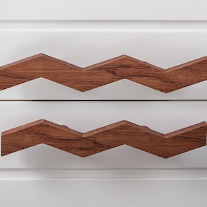 May include: Two brown wooden drawer pulls with a zig-zag design. The pulls are mounted on a white surface.
