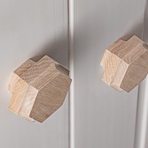 Peut inclure: Deux boutons de meuble géométriques en bois avec une finition marron clair. Les boutons ont un design à facettes et sont montés sur une porte d'armoire gris clair. Les boutons sont un élément de design unique et moderne pour les meubles.
