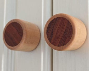May include: Two cylindrical cabinet knobs featuring a light-colored natural wood outer ring and a contrasting dark brown wood circular center inlay. The knobs are mounted on a white vertical paneled surface with a smooth, matte finish.