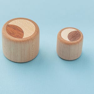 May include: Two wooden cylindrical objects with a light wood base and a contrasting inlaid design on top. The design features a light-colored crescent shape and a darker, reddish-brown accent. The objects are set against a light blue background.