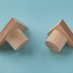 Pode incluir: Dois suportes de prateleira triangulares de madeira com um poste cilíndrico no centro. Os suportes são de cor castanho claro e os postes são de um tom castanho ligeiramente mais escuro.