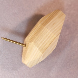 May include: A light-colored wooden knob with a faceted, geometric shape and a visible wood grain pattern. A single gold-toned metal screw is embedded into the side of the knob, extending outward from the faceted surface of the wooden object.