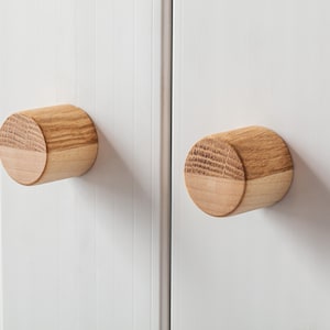 Puede incluir: Dos pomos de armario de madera con un acabado marrón claro. Los pomos son redondos y tienen una superficie ligeramente texturizada.