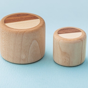 Könnte beinhalten: Zwei zylindrische Holzbehälter, einer groß und einer klein, stehen nebeneinander. Die Oberseiten der Behälter haben ein zweifarbiges Design, mit einem Abschnitt aus hellem Holz und einem Abschnitt aus dunklerem Holz. Die Behälter sind aus hellem Holz gefertigt.